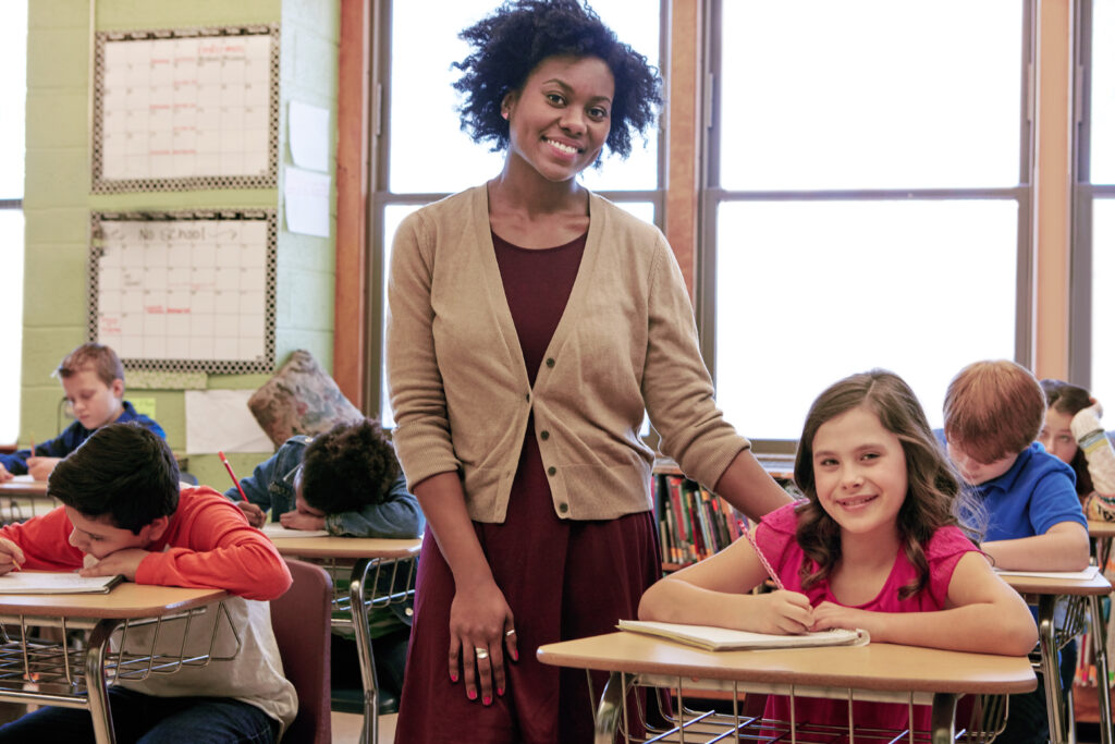 A young woman is standing in the middle of the frame with her left hand on the back of a student's chair. Both are smiling at the camera. The rest of the students are focused on their schoolwork.