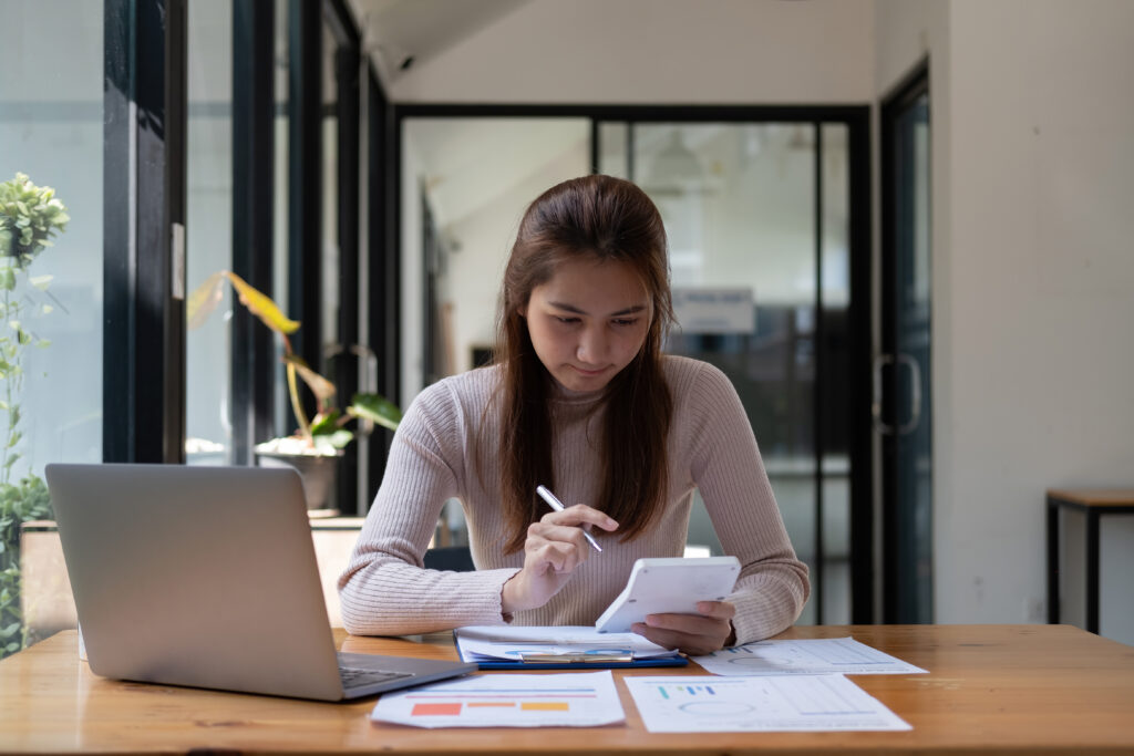 A young woman holding a calculator in her left hand and a pen in her right. She is looking at the calculator and there are lots of papers on the table in front of her as well as a laptop.