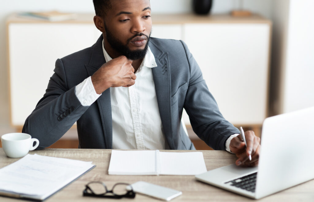 Man looking at his computer. His left hand is holding a pen and is on the trackpad of the computer. His right hand is under his chin.