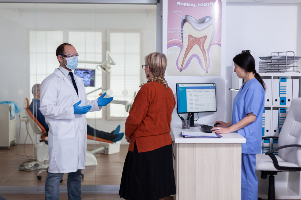 Three people at a dentist office. The dentist on the left is explaining something to a women in the middle. There is a dentist on the right looking at something on the computer.