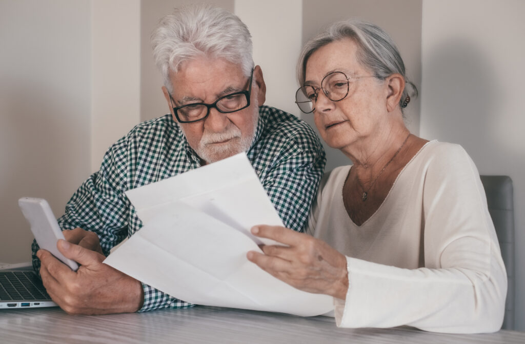 An elderly couple looking at a papers. The man on the left is holding a white phone while the woman on the right is reading off two pieces of paper.