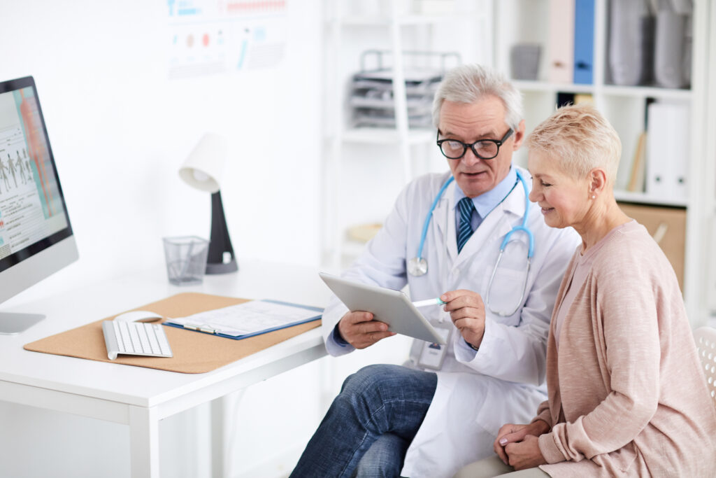 An elderly woman looking at a clipboard that a doctor is holding. They are sitting in a room that appears to be a doctors office.