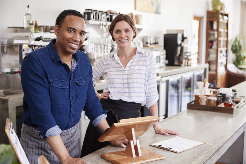 Two people standing behind the counter of a coffee shop. They are both looking into the camera and smiling.