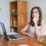 A woman sitting at a desk. Her right arm is rest on the desk and the left arm in on her chin. There is a computer in front of her.