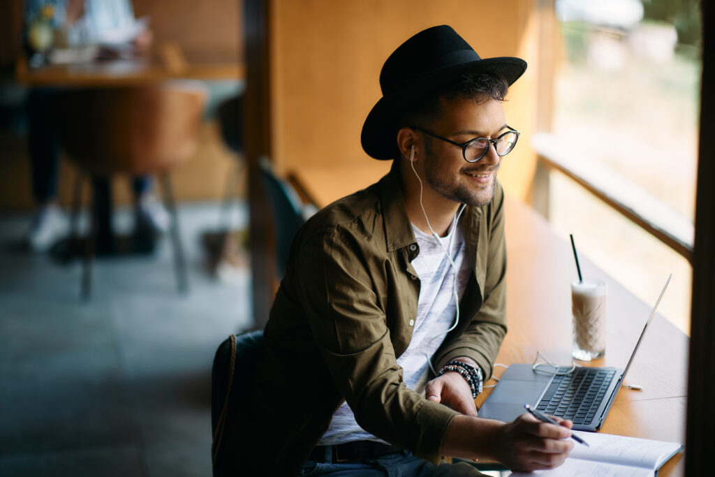 Young man wearing a fedora at a cafe working on a laptop. He has a pen and paper in front of him as well.