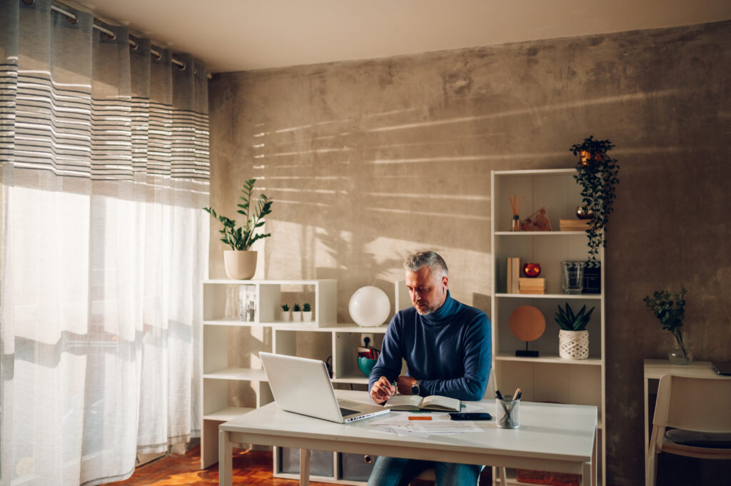 A man sitting at his at home office with a laptop and notebook in front of him. He's holding a pen in his right hand and looks frustrated.