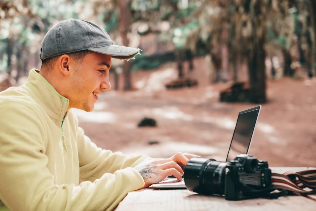 A young man wearing a jean baseball cap and smiling as he's looking at a computer screen. A camera is also on the table he is working on. The background implies that he is outside