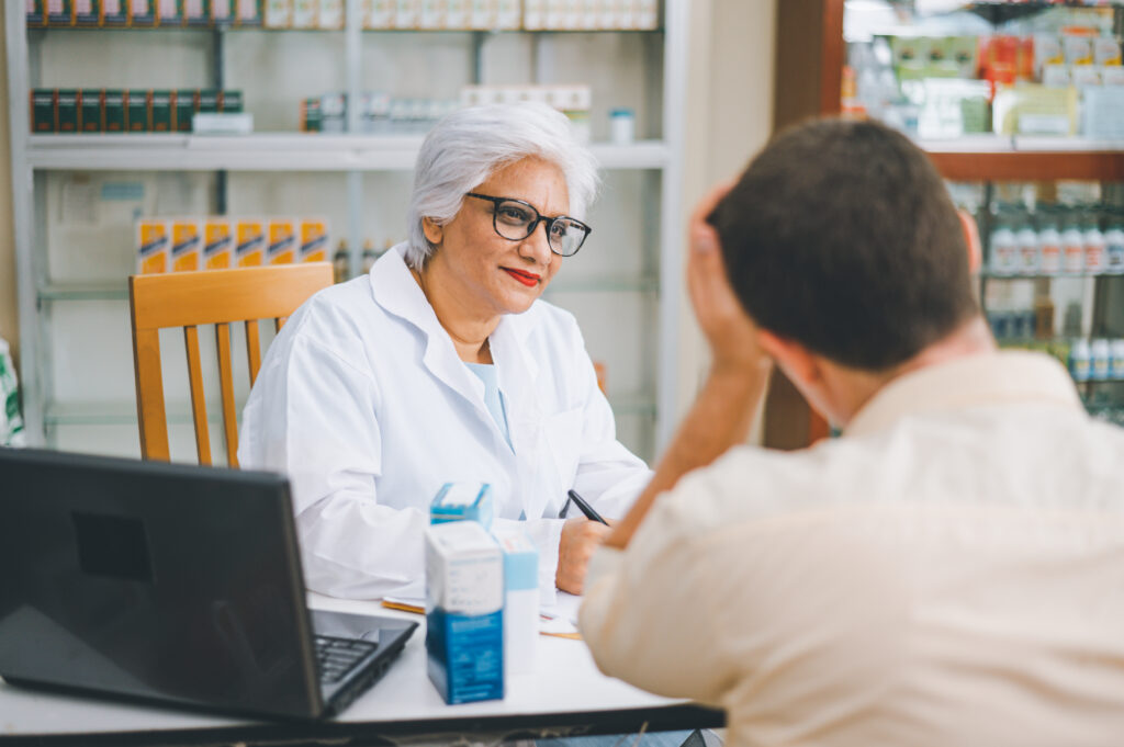 A pharmacist looking at a man whose left hand is holding his head up. There are a few boxes of medicine and a laptop on the desk in front of them.