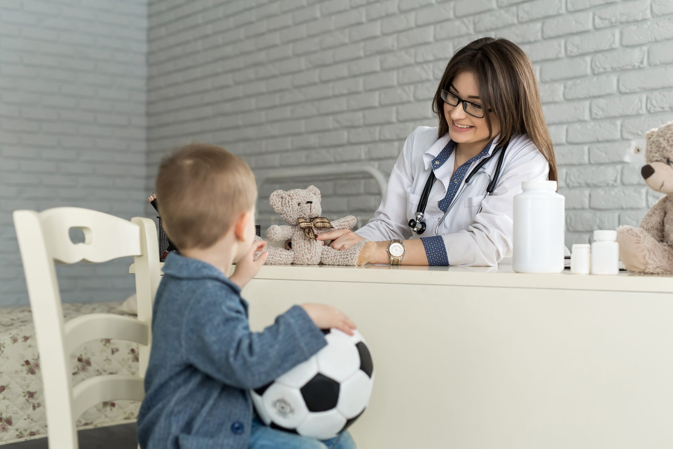 A pediatrician seated at a desk speaking with a seated child holding a soccer ball.